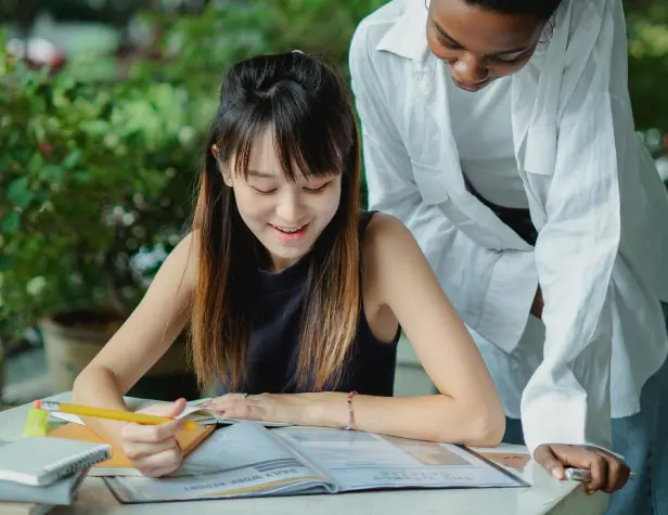 Une jeune fille souriante travaillant de face à une table, ave un professeur particulier en train de l'aider derrière elle lors d'un cours à domicile.