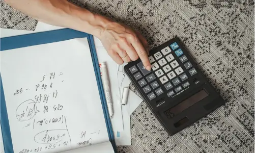 Une vue de haut montrant la main d'un homme en train d'utiliser une calculette, avec une feuille de calcul de mathématiques à côté.